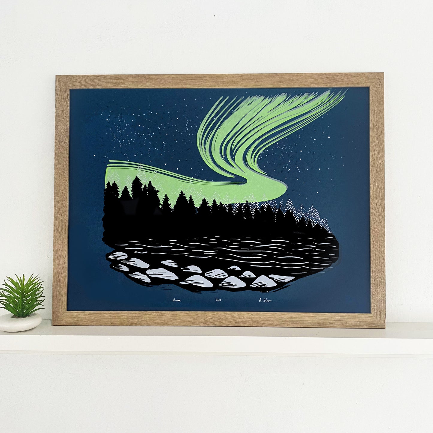 Northern lights artwork. A rich neon green aurora ink on top of a dark blue backround behind the shadows of a Black Forest. A lake in the foreground showing some light on rocks. Framed photo of aurora borealis